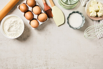Top view of baking ingredients and kitchen utensils forming a border on a light stone background. Eggs, butter, flour, and sugar with ample copy space for a recipe © photoguns