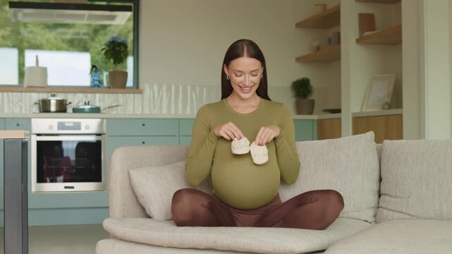 Pregnant woman holding tiny baby shoes over baby bump at home, 4K