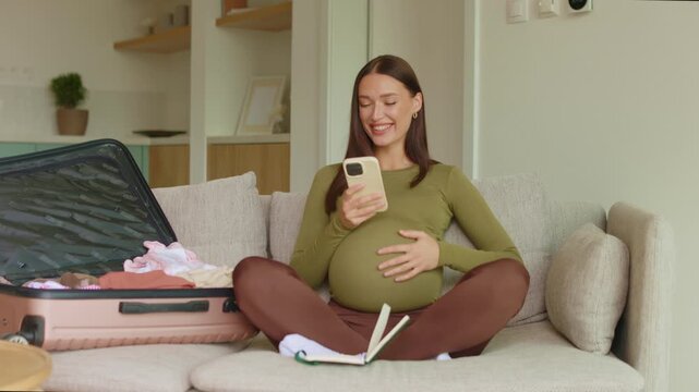 Smiling pregnant woman using smartphone at home and tenderly caressing belly, 4K