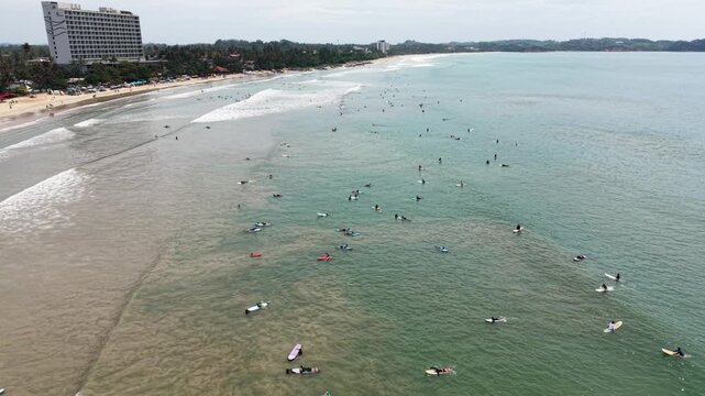 Aerial view of surfers waiting for waves in the turquoise ocean near the sandy coastline and a luxury hotel building in Weligama, Southern Province, Sri Lanka.