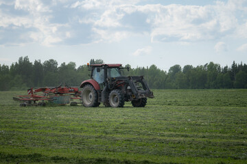 Fototapeta premium Tractor raking hay in green rural farm field under cloudy summer sky