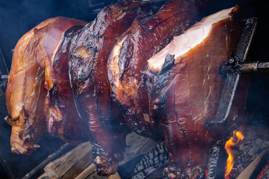 Pork ham on a spit over an open fire against a background of smoke. Close-up of meat slowly rotating on a metal skewer using an electric drive. Concept of popular street food in Europe.