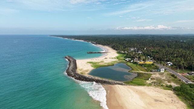 Aerial view of a sandy beach with a stone breakwater, turquoise ocean waves, and lush palm tree forests along the coast in Peraliya, Southern Province, Sri Lanka.