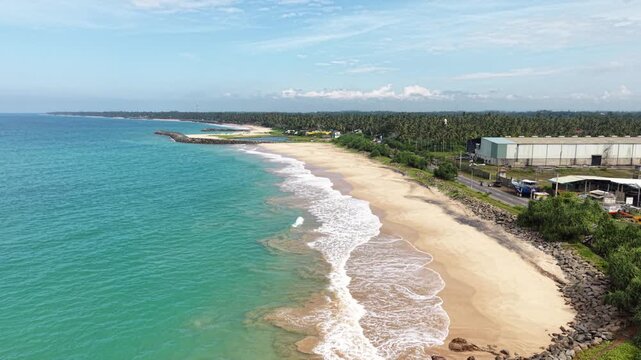 Aerial view of the turquoise coastline with white waves crashing on a sandy beach lined with palm trees and industrial buildings in Peraliya, Southern Province, Sri Lanka.