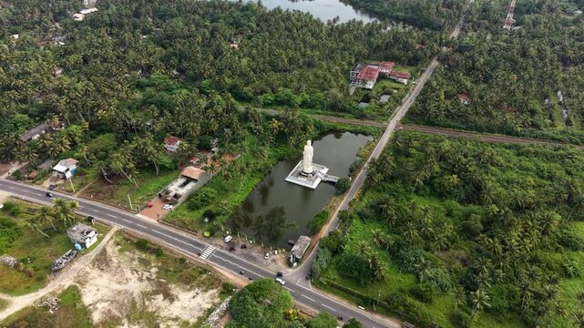 Aerial view of Tsunami Honganji Viharaya standing in a pond surrounded by lush palm trees and a coastal road Peraliya, Southern Province, Sri Lanka.
