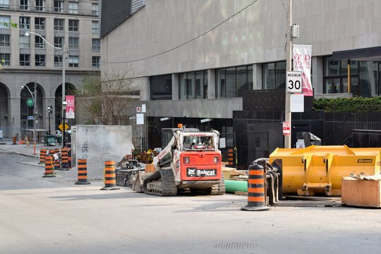 Urban road construction site in downtown Toronto featuring a compact track loader, traffic barrels, construction materials, and street infrastructure improvement work, May 2026