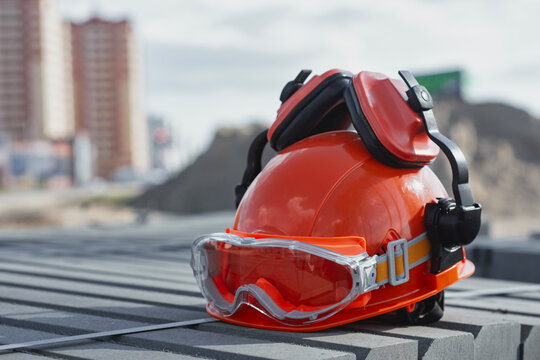 Orange construction hard hat with safety goggles and black earmuffs resting on concrete surface. Personal protective equipment, occupational safety and worker protection at construction sites.