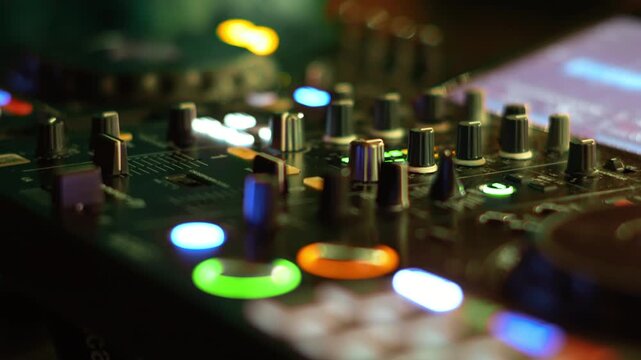 Close perspective of a professional dj controller showing rows of knobs faders and a glowing cue button surrounded by colorful club lighting