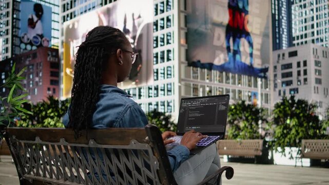 Writing code on laptop, implementing programming language, software developer on park bench. Black girl developing software interface, freelancing outdoors for productivity. City life.