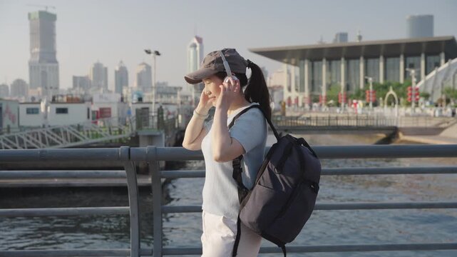 A smiling young female traveler wearing casual summer clothing and a backpack stands by a waterfront railing, waving at the camera with a city skyline in the background.