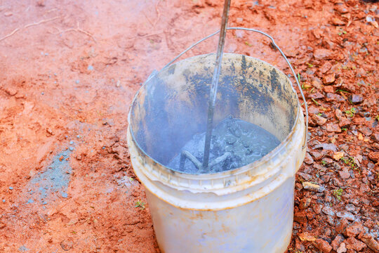 Worker stirs concrete mix in bucket at construction site with tools on dirt surface.