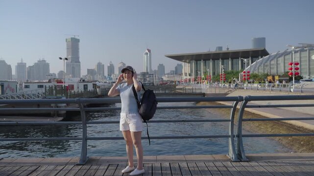 A young female jogger wearing casual sportswear and headphones stops along a wooden waterfront promenade overlooking a city skyline on a sunny day.