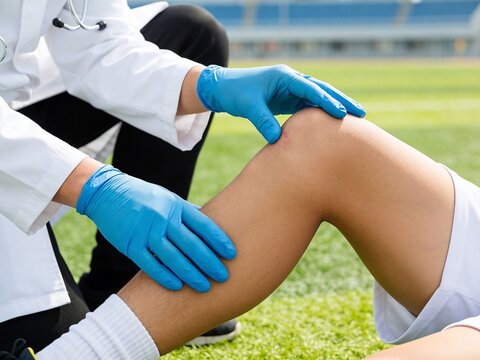 A healthcare professional in a white coat examines a knee injury on a person at a sports field.