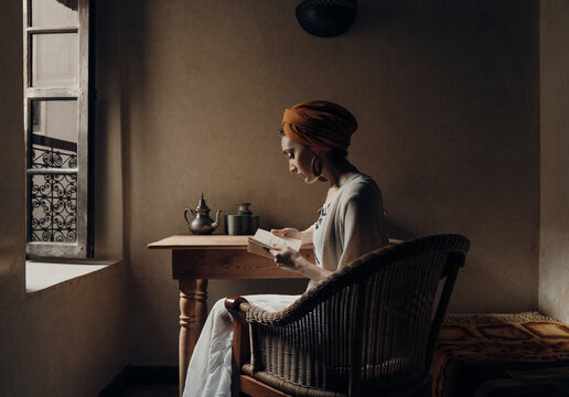 Stylish woman in turban reading in armchair near open window
