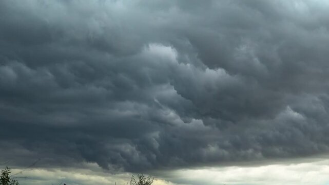 Layers of dark grey storm clouds looming over the silhouette of trees with a faint pale glow near the horizon before rainfall