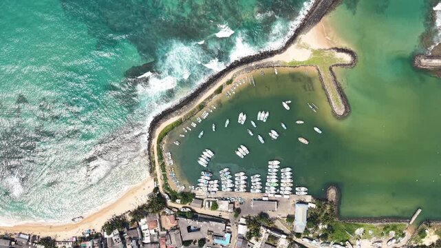 Aerial view of white fishing boats moored in a protected harbor next to crashing waves on a sandy beach in Hikkaduwa, Southern Province, Sri Lanka.