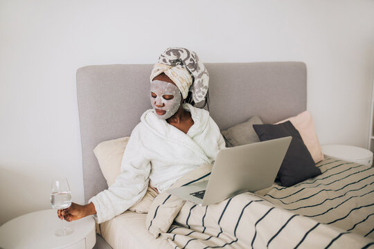 African american woman with face mask sitting with laptop in bed