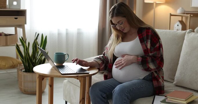 Pregnant woman working with laptop and touching her belly on sofa at home. Camera moving forward