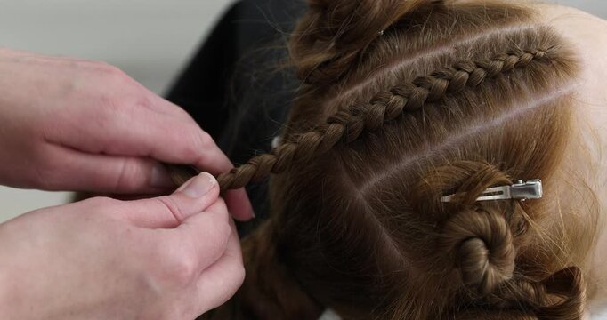Hairstylist braiding teenage girl's hair indoors, closeup