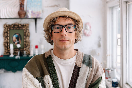 Man with curly hair wearing glasses and a hat stands in a art-room