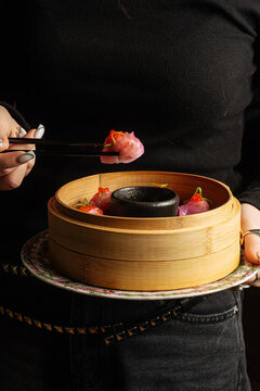 Person holding bamboo steamer with pink dumpling in chopsticks