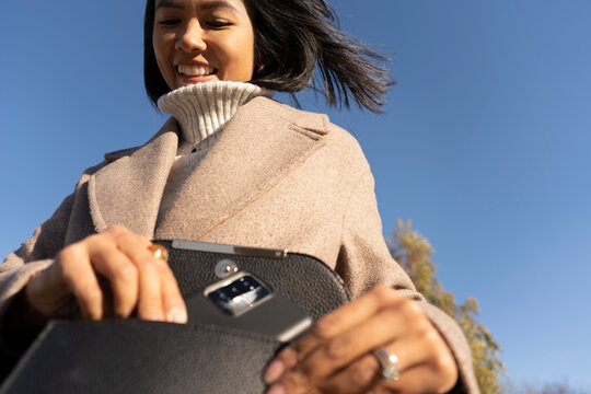 Woman Smiles While Taking a Mobile Phone From Her Purse Under Blue Sky