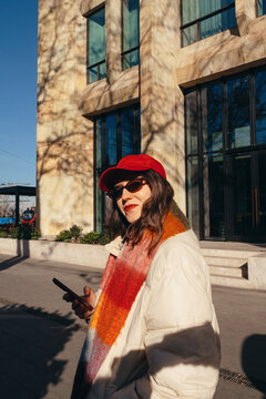 Stylish woman in red cap checking phone in sunlit urban setting