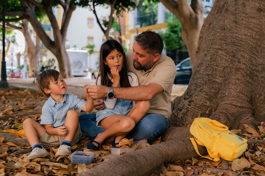 Children Share Snacks With Father Under a Large Tree in Park