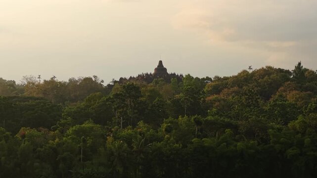 Aerial view of Borobudur Temple peaking over a dense green forest canopy under a hazy golden sky in Kecamatan Borobudur, Jawa Tengah, Indonesia.