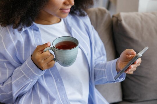 Obscure face of cropped black woman with tea using smartphone on sofa