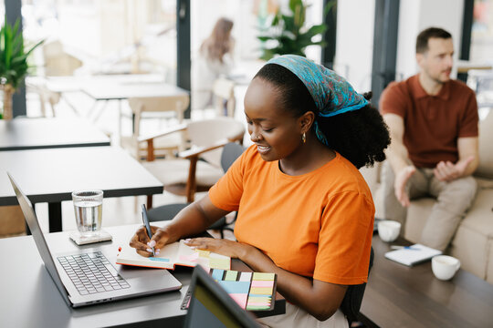 Woman Writing Notes While Using Laptop in Cafe