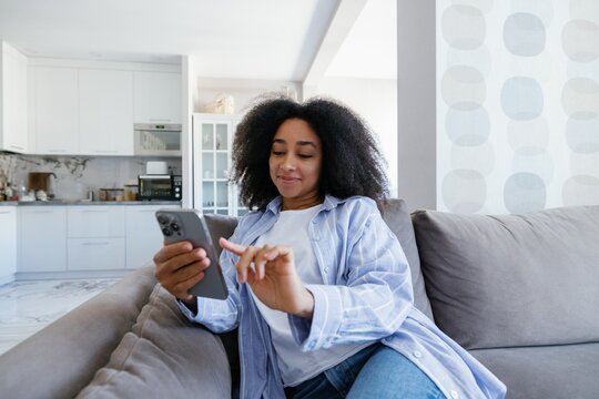 Young smiling black woman using and pushing on mobile phone on sofa