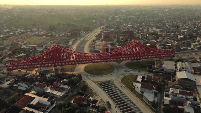Aerial view of Jembatan Kereta Api Joglo, a red steel truss bridge spanning over a busy urban roundabout during golden hour in Kecamatan Banjarsari, Jawa Tengah, Indonesia.
