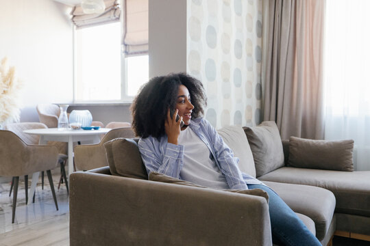 Young smiling black woman talking on smartphone on sofa at living room