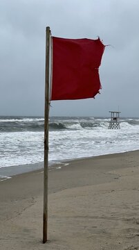Red warning flag on a wooden pole stands on a windy beach, rough waves roll under a gray sky, distant lifeguard tower suggests hazardous surf