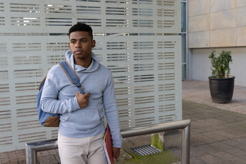 African American teen male standing by metal railing in plaza, holding blue backpack, red folder © wavebreak3
