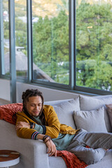 Fototapeta premium African American man reclining on sofa in living room, wearing mustard jacket, showing smartwatch