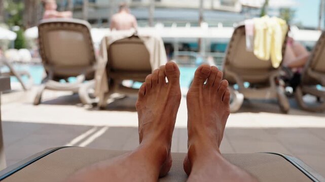 First person view of relaxed feet on sun lounger near swimming pool at sunny resort. Peaceful summer holiday, leisure travel and vacation lifestyle