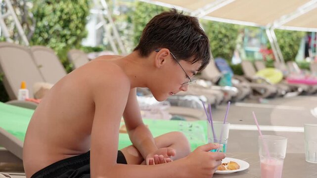 Side view of teenage vacationer eating casual poolside meal with cold drinks at sunny resort. Family summer holiday, leisure dining and tropical travel lifestyle. Lunch time at pool side on vacation