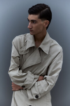 Young man in trench coat standing in profile against grey wall