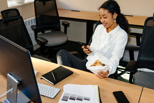 Businesswoman Checking Smartphone While Holding Sandwich at Desk