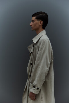 Young man in trench coat standing in profile against grey wall
