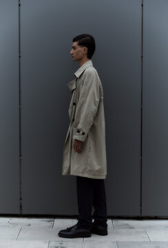 Young man in trench coat standing in profile against grey wall