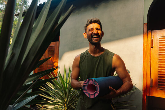 Man Smiles and Holds Yoga Mat in Outdoor Space With Plants