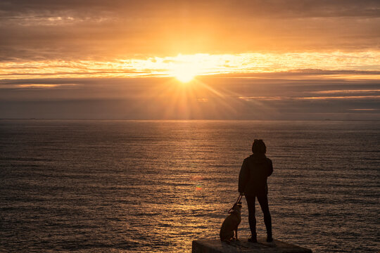 Silhouettes of Woman and Dog Watching Sunset on Galician Cliffs
