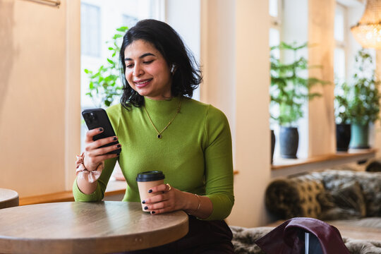 Smiling woman using smartphone and drinking coffee in a bright cafe