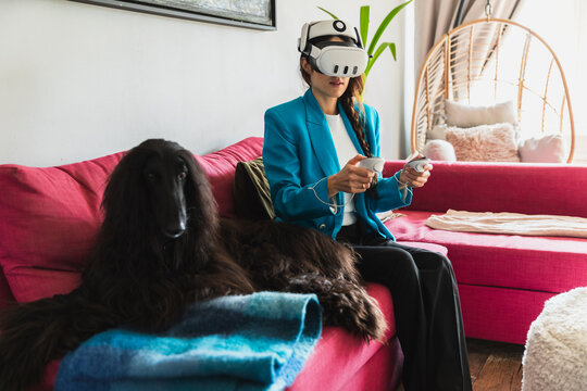 Woman using a VR headset on a pink sofa next to her sleeping dog