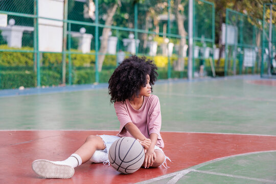 Girl with basketball sits on outdoor court, looking away.