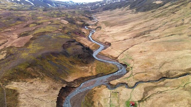 Aerial view of a winding river flowing through the rugged Brunavik valley landscape with snow-capped mountains under a cloudy sky in Bakkagerdi, Mulathing, Iceland.