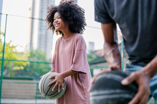 Smiling girl with basketball, father nearby on outdoor court.
